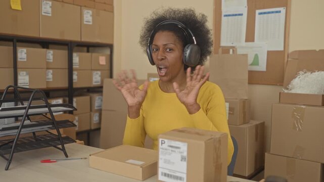 Woman wearing headphones holding a parcel and pointing finger to shipping label in a packing building with stacked boxes; small business pride.
