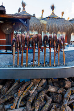 Traditional Spanish Espeto de Pulpo. Grilled octopus skewers cooking over charcoal fire at a beach chiringuito in Malaga, Andalusia. Authentic Mediterranean seafood gastronomy.