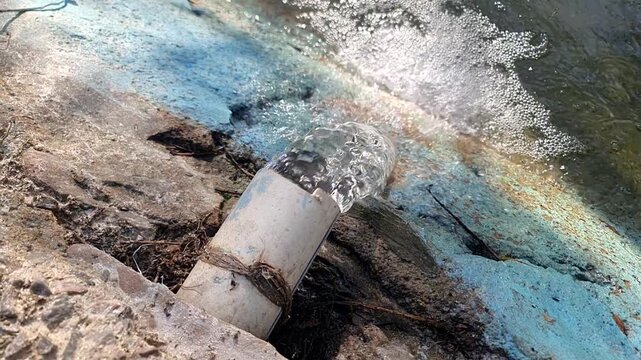 Water flowing out of a white plastic pipe into a drain with bubbles and sunlight.