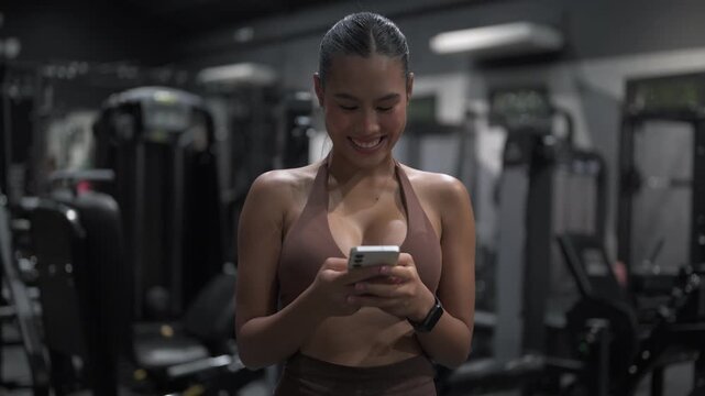 Portrait of a young, fit happy Thai woman in a modern gym exercising texting and using smartphone
