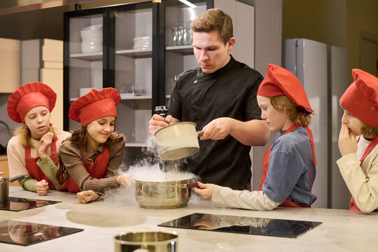 Young adult male chef guiding children preparing food during cooking class, demonstrating kitchen skills and teamwork. Useful for culinary education, kids workshops, parenting campaigns