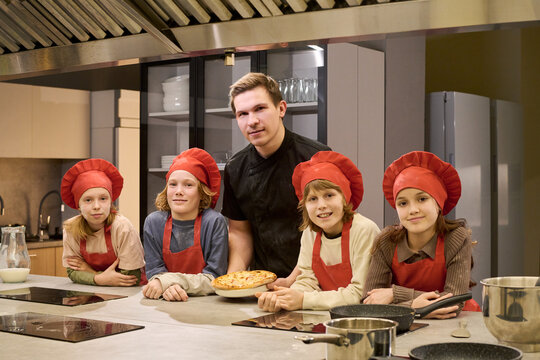 Portrait of Caucasian male chef guiding school age kids in cooking class, presenting baked pizza in training kitchen. Useful for culinary education, teamwork, youth workshop promotion