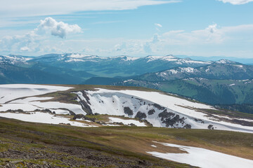 Scenic alpine landscape with sunlit snowy field on stony hill with view to big mountain range with forest and snow far away under cloudy sky. Stone outcrops among snows in sunlight in high mountains. © Daniil
