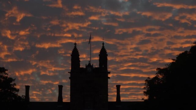Wide shot Silhouette of gothic university building against dramatic orange sunset clouds with flag gently moving, trees either side of frame