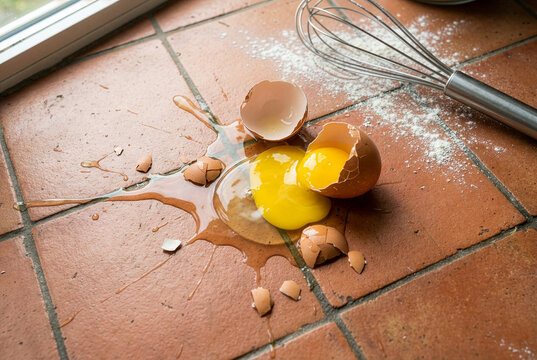 Broken raw egg splashed on a terracotta kitchen floor next to a whisk and spilled flour