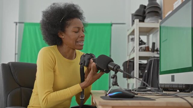 Woman adjusts camera lens and holds strap while seated at desk with microphone in studio; focused creativity.