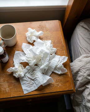 Pile of crumpled tissues, an empty mug, and medicine on a wooden bedroom nightstand during a sick day
