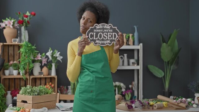 Woman florist in green apron holding 'closed' sign and pointing finger among flowers on table in building; quiet resignation.