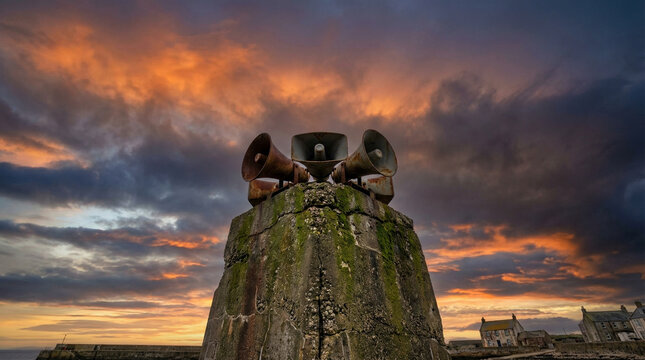 Dramatic coastal scene with a historic tsunami warning siren silhouetted against a vibrant sunset sky, surrounded by rugged stone structures and distant village rooftops