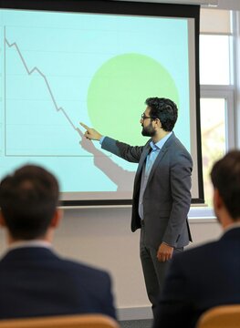 Man Standing In Front Of Audience Presenting Information On Projection Screen Behind Presenter Dressed Semi Formally With Dark Blazer Lighter Colored Shirt Complemented