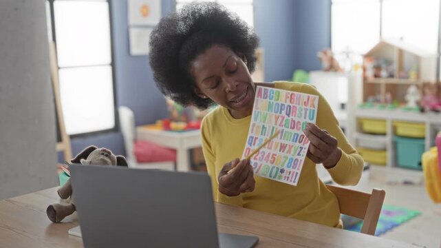 Woman pointing with a pencil at a colorful alphabet chart during a laptop lesson in a classroom; learning engagement.