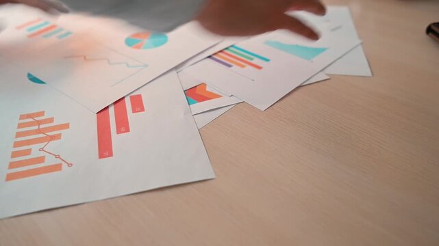Hands organize and sort various papers filled with charts and graphs on a wooden desk. Different types of data presentations are observed, showing detailed information on various topics