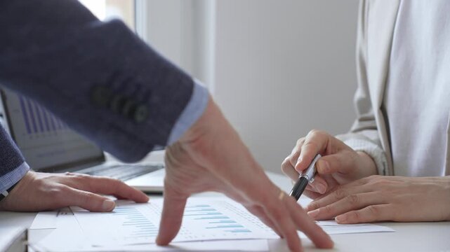 Financial analysts reviewing laptop screen and printed documents, pointing at data charts and discussing investment strategies together in modern office setting. Business people concept