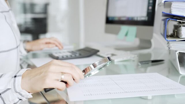 Businesswoman hands analyzing financial document with magnifying glass, working on computer in office, reviewing data. Audit and taxes in business