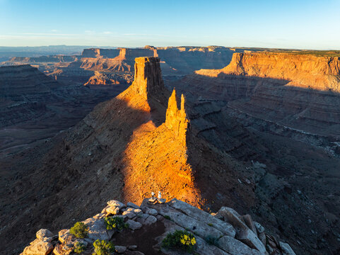 Aerial view of Canyonlands National Park featuring dramatic red rock buttes and spires illuminated by golden hour light with two people standing on a rocky ridge Utah, United States.