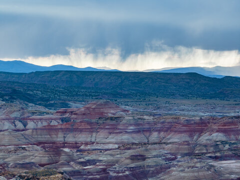 Aerial view of the colorful banded rock formations of the Bentonite Hills under a dramatic sky with rain shafts in Wayne County, Utah, United States.