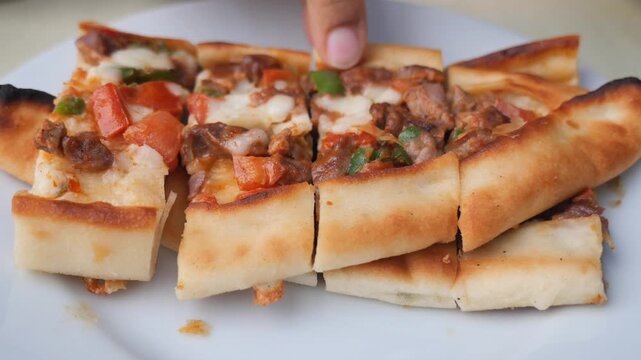 A hand lifts a slice of turkish pide with melted cheese and meat from a white plate.