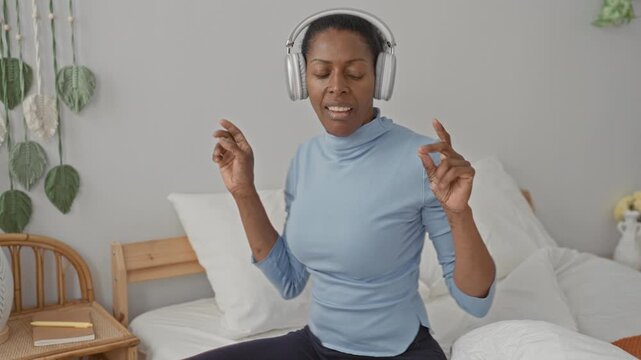 Woman with headphones on bed dancing with hands raised and eyes closed in studio; joy relaxation calm.
