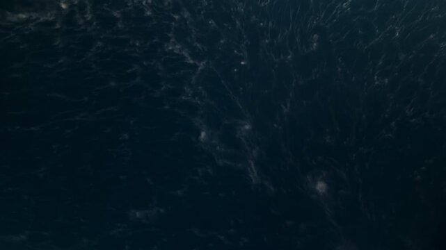 Aerial top down view of dark deep ocean water surface with scattered white foam patterns and wave ripples. Moody atmospheric seascape showing turbulent water texture and natural movement.