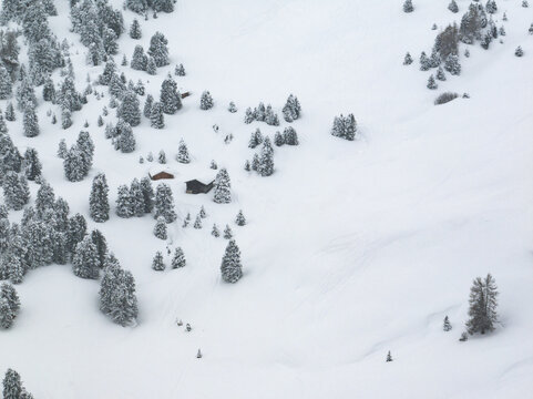 Aerial view of the snow-covered Sella Pass with small wooden huts nestled among pine trees on a winter slope in Selva, Trentino-South Tyrol, Italy.
