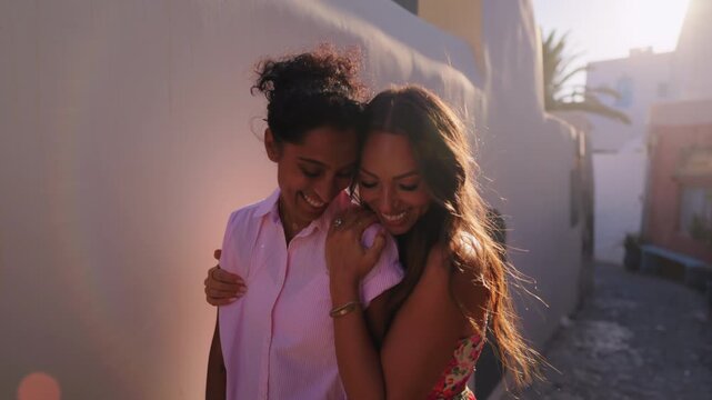 Same sex couple shares intimate moment on Santorini island in Greece. Indian and diverse lesbians embrace during holiday with warm golden sunlight. LGBTQ love and connection outdoors.