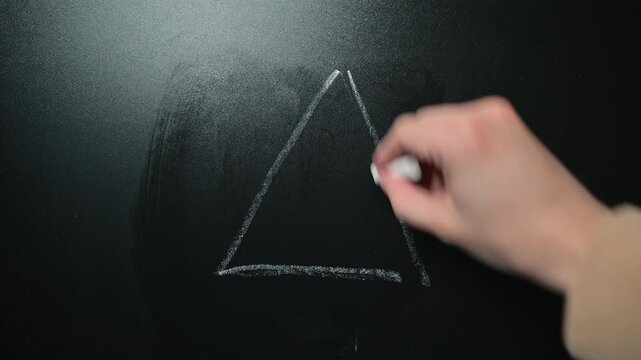 A hand draws a triangle shape using chalk on a dark surface in a well-lit area.