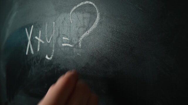 A hand writes mathematical symbols on a blackboard using white chalk, focusing on equations and variables.