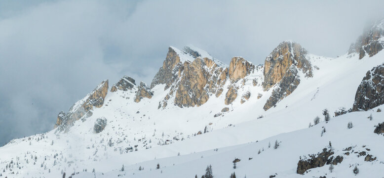Aerial view of the jagged snowy peaks of Passo Giau with misty clouds and sparse pine trees on the slopes in Colle Santa Lucia, Veneto, Italy.