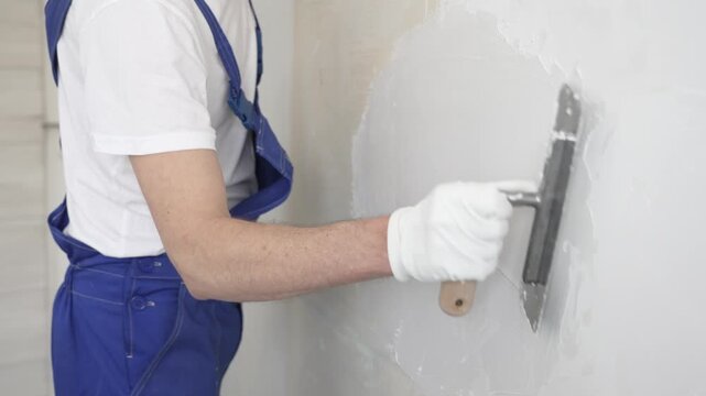 Unknown construction worker man plastering white wall, wearing blue overalls and protective white gloves during apartment renovation project, close-up