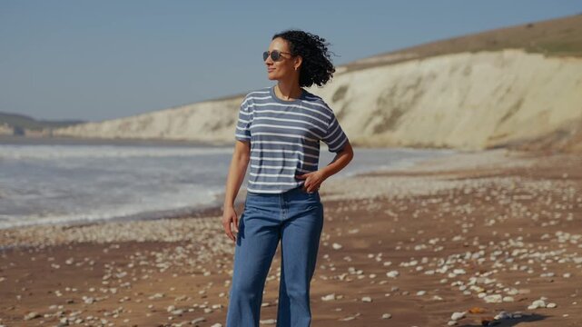 Woman smiles, phone to ear, walking seaside. She's dressed in striped top, jeans, sunglasses. Beach stretches wide with shells, waves, cliffs. Sunlight glows warm, clear skies above