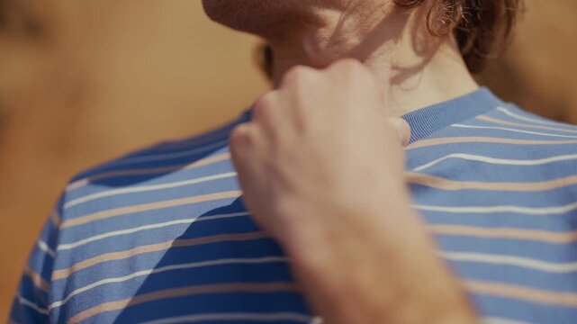 Man in striped shirt touches his neck. Hands show thoughtful or nervous gesture. Warm, golden light bathes his skin. Sunlit outdoor setting suggests calm, personal moment