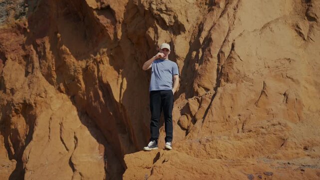 Man stands on rocky ledge, sunlit, calm. He adjusts cap, looks around, relaxed posture. Bright orange rock walls frame his presence, natural. Ideal for travel, adventure, or outdoor lifestyle content