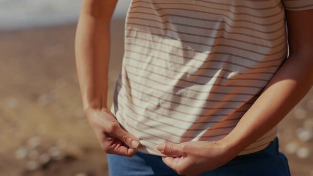 Person adjusts loose shirt near waist. Hands grip fabric, suggesting discomfort or looseness. Sunlit beach setting implies casual, outdoor context. Ideal for lifestyle, fitness