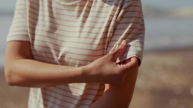 Sunlit beach, person in striped shirt. Hands gently pull sleeve to expose arm. Soft focus, warm tones, calm moment. Relaxing, casual, summertime vibe. Ideal for wellness, travel, or lifestyle content