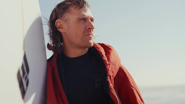 Surfer leans against board, gaze fixed on horizon. Red jacket contrasts with bright sky and calm sea. He appears thoughtful, waiting for the next wave