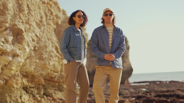 Sun-drenched cliffs meet calm sea waves. Two people stroll, smiling, barefoot, wearing casual clothes. One adjusts sunglasses, the other arms crossed. Warm, golden light bathes the rocky shore