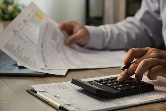 Close up of businessman using calculator and reviewing financial documents on desk, analyzing business report, accounting data, and tax paperwork in modern office, representing finance, audit, and bud