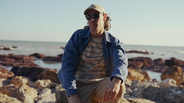 Man sits on rocky shore, gazing out at sea. He adjusts sunglasses, then looks ahead with calm focus. Bright sun glints off water, casting warm tones across scene. Perfect for travel, vacation