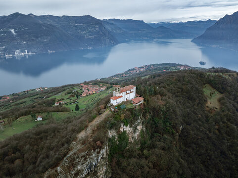 Aerial view of Lake Iseo with the Santuario della Madonna della Ceriola on a rocky peak surrounded by dense forests and mountains under a cloudy sky Lombardy, Italy.