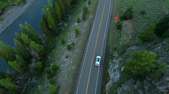 Drone footage follows a white car traveling along Beartooth Highway in Montana through lush green forest with a winding river below. Pine trees line the scenic mountain road during twilight hours.