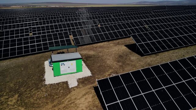Aerial view of solar power station with rows of solar panels and green maintenance building on dry land under clear blue sky in a renewable energy field