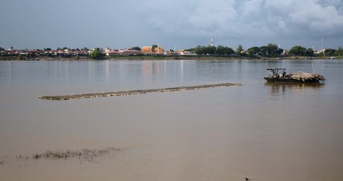 wooden boat towing the piece of the structure of the new bamboo bridge on the Mekong river , with the Kampong Cham town and temples visible on the opposite bank.