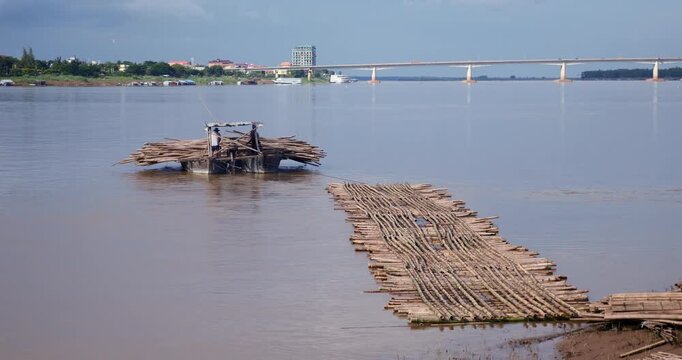  wooden boat towing the piece of the structure of the new bamboo bridge on the Mekong River in Kampong Cham with the Kizuna Bridge in the background. 