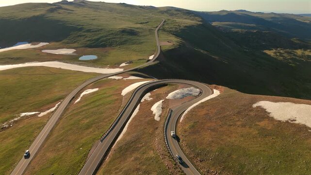 Beartooth Highway in Montana winds through alpine terrain with dramatic hairpin curves. Cars travel along the scenic mountain pass with patches of snow and green meadows visible from above.