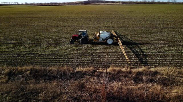 Red tractor with white sprayer attachment moves across green agricultural field, spraying crops while shadows stretch across the soil under a clear blue sky