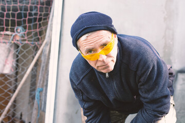 Professional Middle-Aged Worker Wearing Yellow Safety Glasses and Blue Beanie in Outdoor Workshop Setting © dizfoto1973