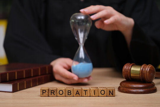 A judge holds an hourglass filled with blue sand, symbolizing time in a courtroom setting. Nearby, wooden gavel and stacked legal books represent the legal process and decisions being made.