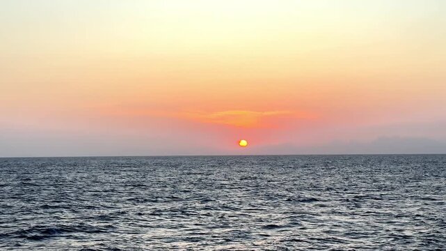 Vibrant crimson sun dips below the horizon of the vast open ocean during a serene summer evening captured from a low sea level perspective with soft hazy clouds and golden light across the water