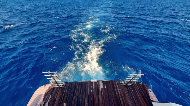 A serene high angle view of the teak wood deck of a luxury yacht cruising through deep blue Mediterranean waters on a sunny summer day creating a vibrant white sea foam wake under a clear sky