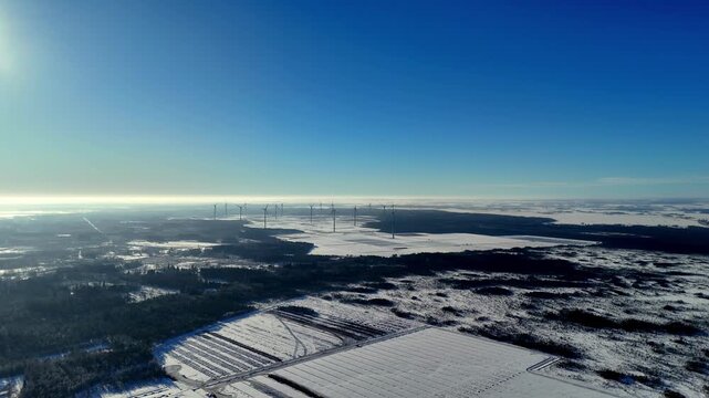 Winter landscape aerial view of vast snowy fields and dark evergreen forests under a bright clear blue sky with a wind farm on the horizon during a cold sunny day captured from a high altitude drone